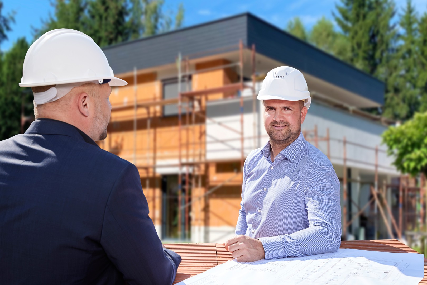 Zwei M&auml;nner tragen wei&szlig;e Helme und sprechen vor einer Baustelle mit einem Haus im Hintergrund, daneben liegen Pl&auml;ne.