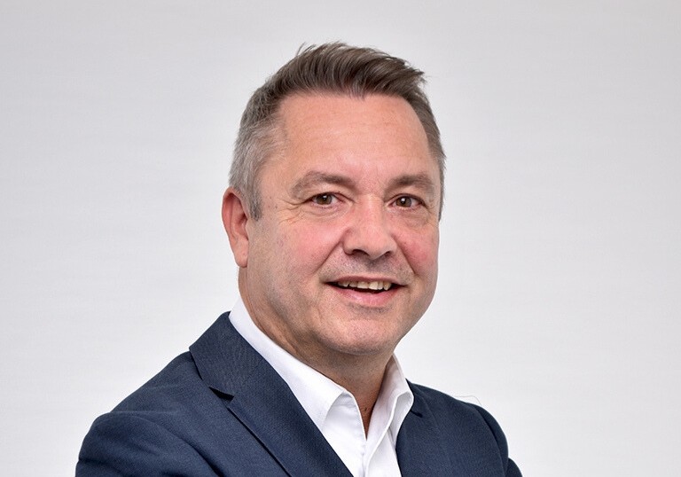 Eine Person in einem dunkelblauen Anzug mit weißem Hemd vor einem neutralen Hintergrund. Das Gesicht ist durch einen Balken verdeckt.