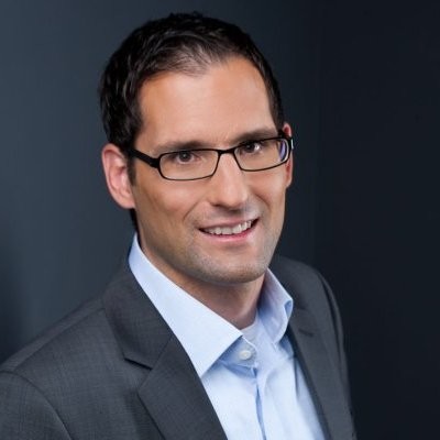 Professionelles Porträt eines lächelnden Mannes mit Brille in grauem Sakko und hellem Hemd vor dunklem Hintergrund. Das Bild wirkt wie ein Business- oder Bewerbungsfoto.