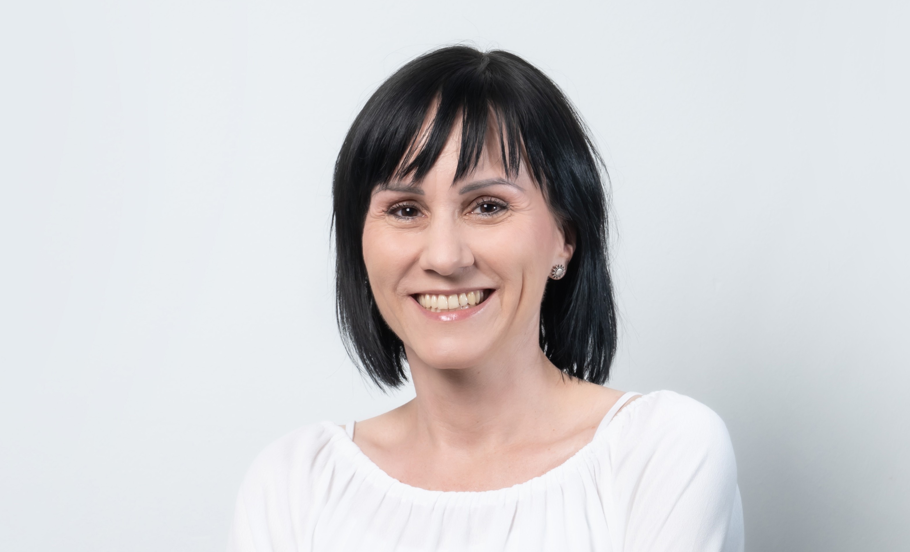 Porträt einer lächelnden Frau mit kurzen dunklen Haaren vor hellem grauem Hintergrund. Sie trägt eine weiße Bluse und kleine Ohrringe, das Bild wirkt freundlich, ruhig und professionell.