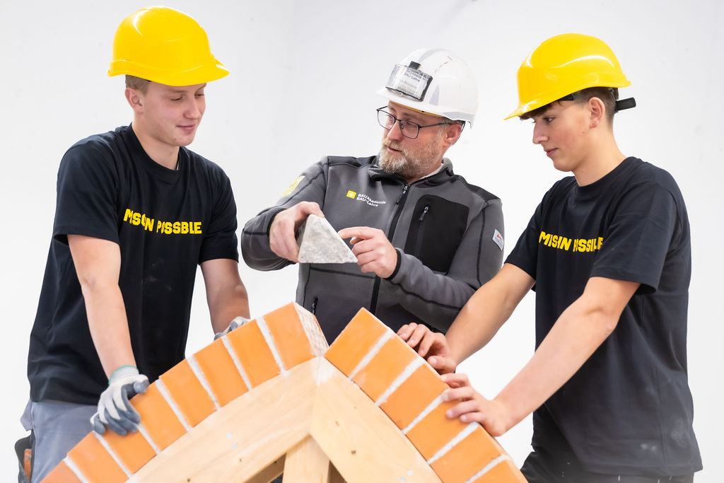 Drei Personen tragen Schutzhelme und arbeiten gemeinsam an einer Architekturkonstruktion aus Ziegelsteinen. Eine Person erklärt oder zeigt den anderen etwas.