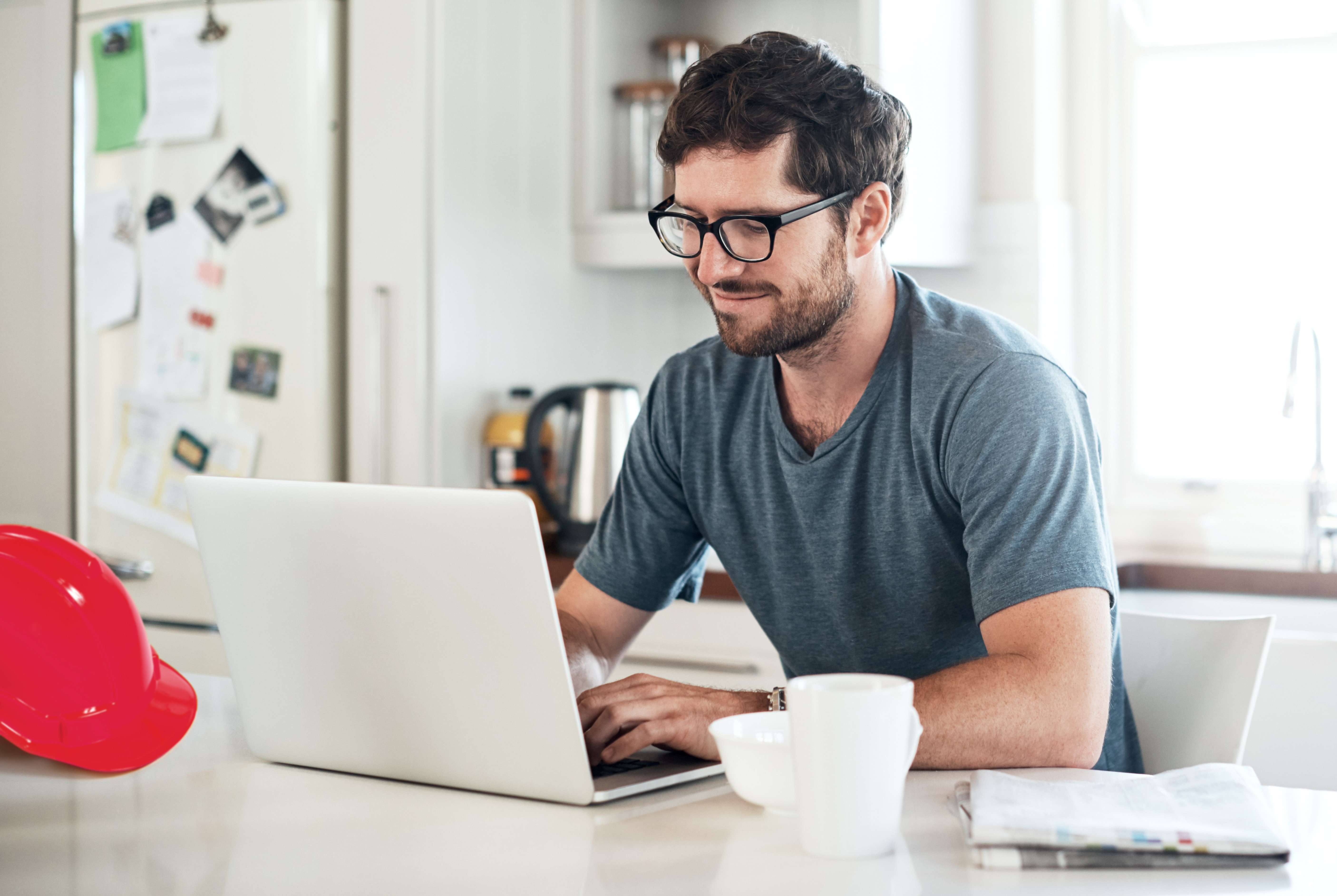 Ein Mann sitzt in einer modernen Küche, arbeitet an einem Laptop und hat eine Kaffeetasse neben sich. Im Hintergrund sind Haushaltsgeräte, ein Kühlschrank und ein roter Schutzhelm zu sehen.