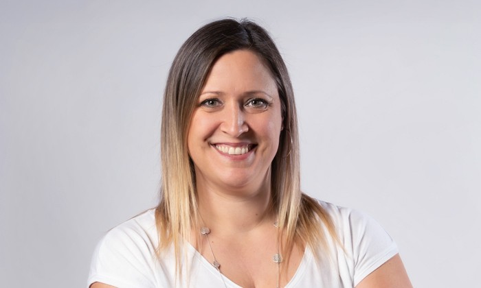 Das Bild zeigt eine Frau in einer weißen Kleidung mit mittellangem Haar, deren Gesicht aus Datenschutzgründen unscharf gemacht wurde. Sie trägt eine Kette und ist vor einem neutralen grauen Hintergrund abgebildet.