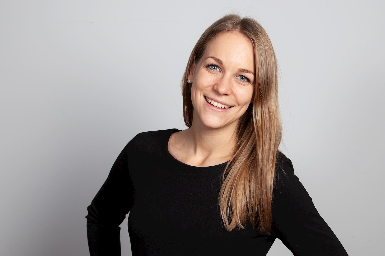 Eine Person mit langen hellbraunen Haaren trägt ein schwarzes Oberteil und posiert vor einem neutralen grauen Hintergrund. Das Gesicht ist unscharf/durch einen Filter verdeckt.