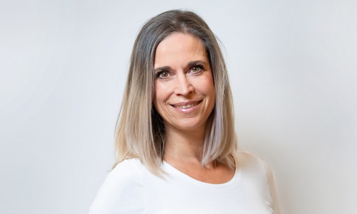 Eine Frau in heller Kleidung steht vor einem neutralen weißen Hintergrund. Ihr Gesicht ist unkenntlich gemacht, sie hat schulterlanges blondes Haar.