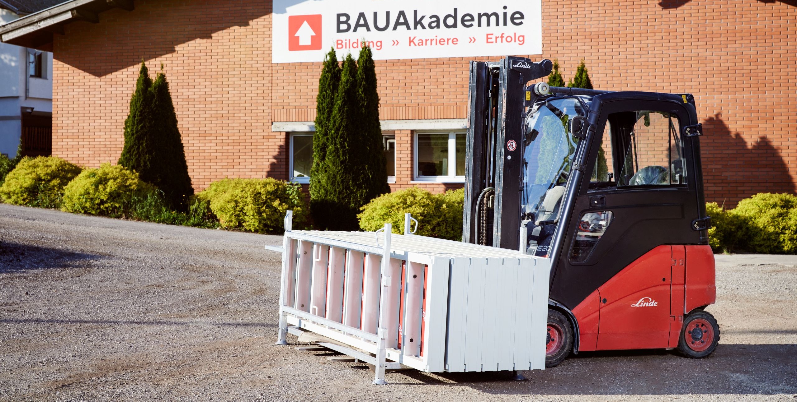 Ein roter Gabelstapler der Marke Linde transportiert eine weiße Konstruktion vor einem Gebäude mit der Aufschrift "BAUAkademie". Im Hintergrund sind Büsche, eine Backsteinfassade und ein Schild zu sehen.