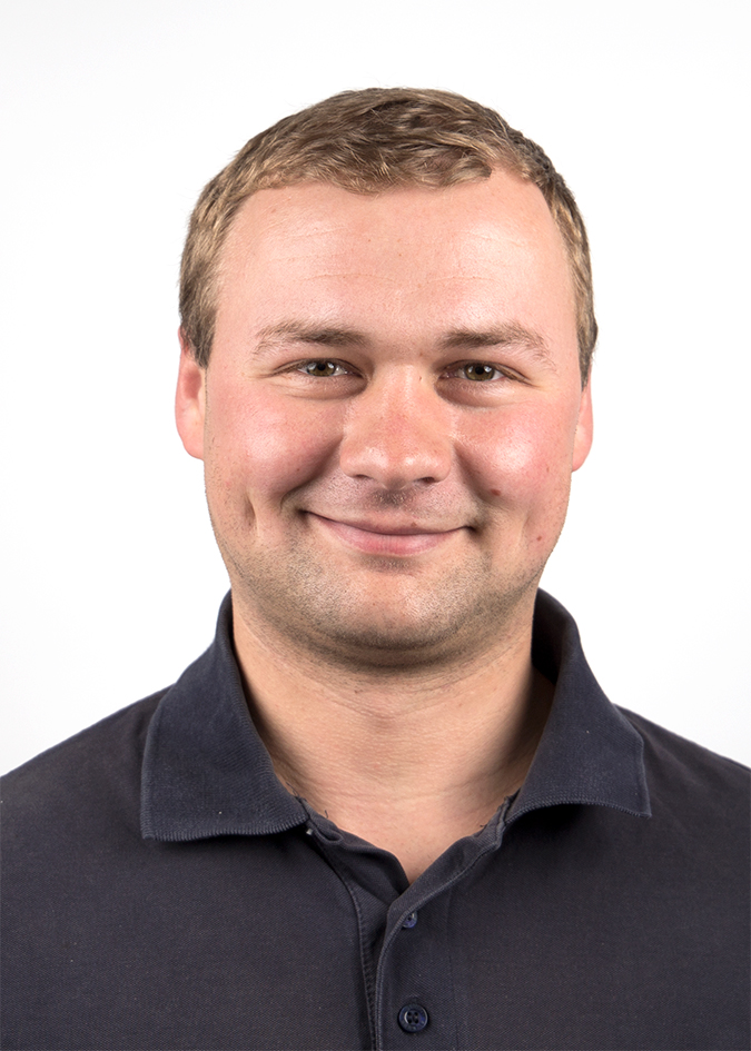 Das Bild zeigt einen Mann in einem dunkelblauen Poloshirt mit einem verschwommenen Gesicht. Der Hintergrund ist weiß.