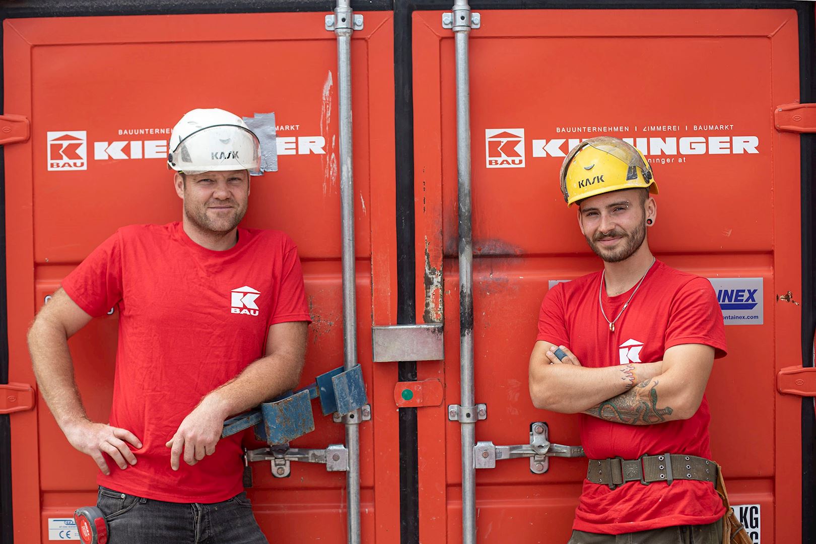Zwei Bauarbeiter stehen vor einem roten Container mit Firmenlogo und tragen rote T-Shirts und Schutzhelme. Einer lehnt lässig, der andere steht mit verschränkten Armen.