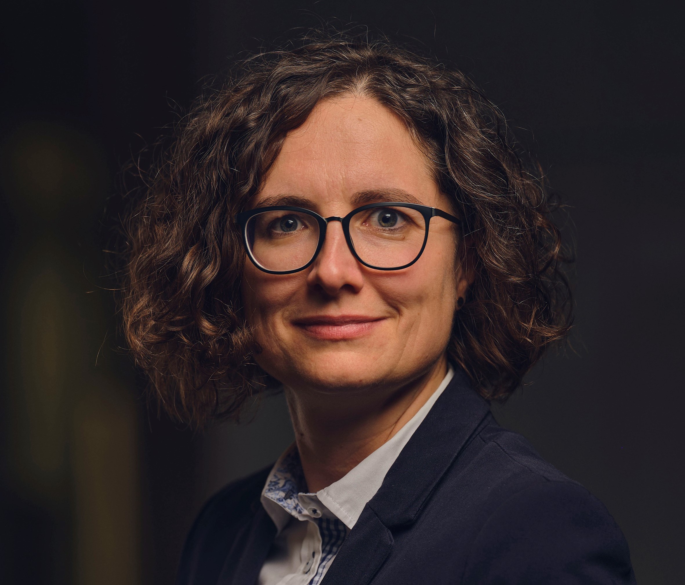 Ein Portrait einer Person mit dunklem, welligem Haar, einem dunklen Blazer und heller Bluse auf dunklem Hintergrund. Das Gesicht ist verpixelt, wodurch keine genaue Identifizierung möglich ist.