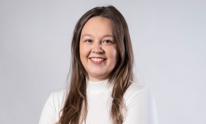 Das Bild zeigt eine Person mit langen braunen Haaren, die ein weißes Oberteil trägt. Das Gesicht ist unkenntlich gemacht und der Hintergrund ist neutral grau gehalten.