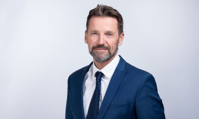 Das Bild zeigt eine Person in einem eleganten blauen Anzug mit Krawatte vor einem neutralen Hintergrund. Das Gesicht ist unkenntlich gemacht.