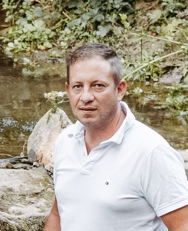 Ein Mann trägt ein weißes Polohemd und steht vor einem natürlichen Hintergrund mit einem Fluss, Steinen und grüner Vegetation. Das Gesicht ist verpixelt.