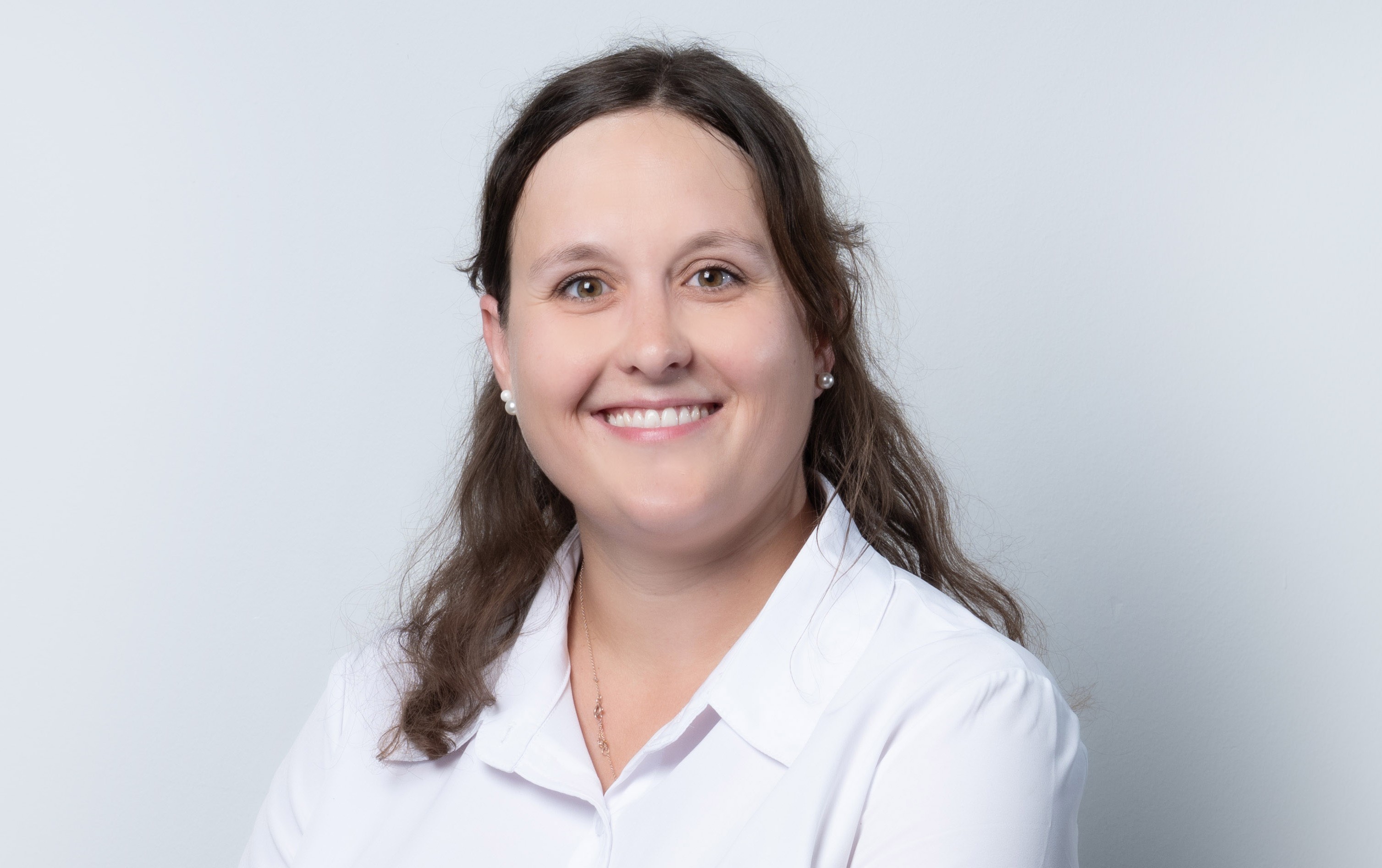 Porträt einer lächelnden Frau mit langen dunklen Haaren vor hellem Hintergrund. Sie trägt eine weiße Bluse, kleine Ohrringe und eine feine Kette. Das Bild wirkt freundlich, professionell und gepflegt.