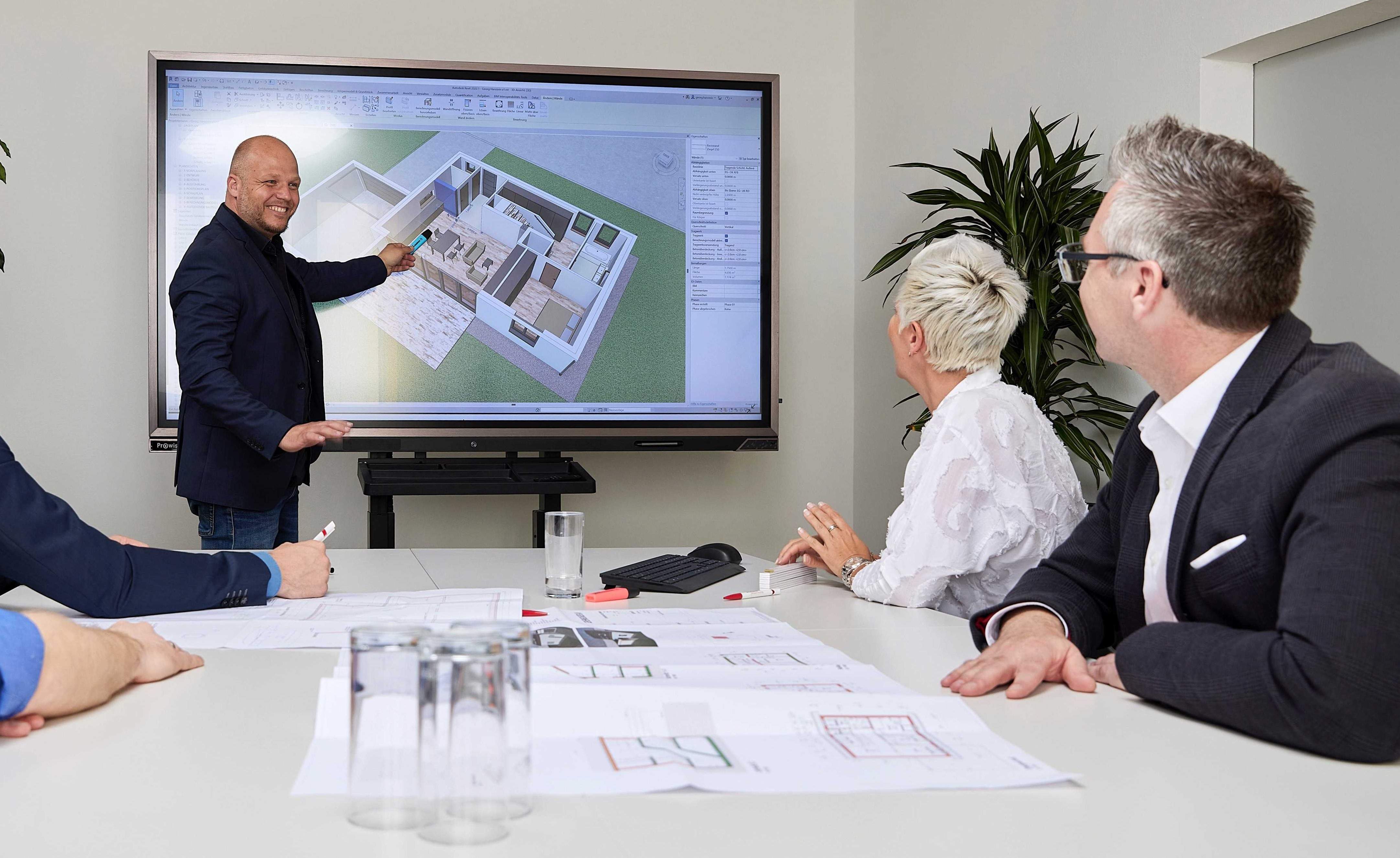 Menschen in einer Besprechung diskutieren über Architekturmodelle auf einem Bildschirm. Baupläne liegen auf dem Tisch. Der Raum ist modern eingerichtet.