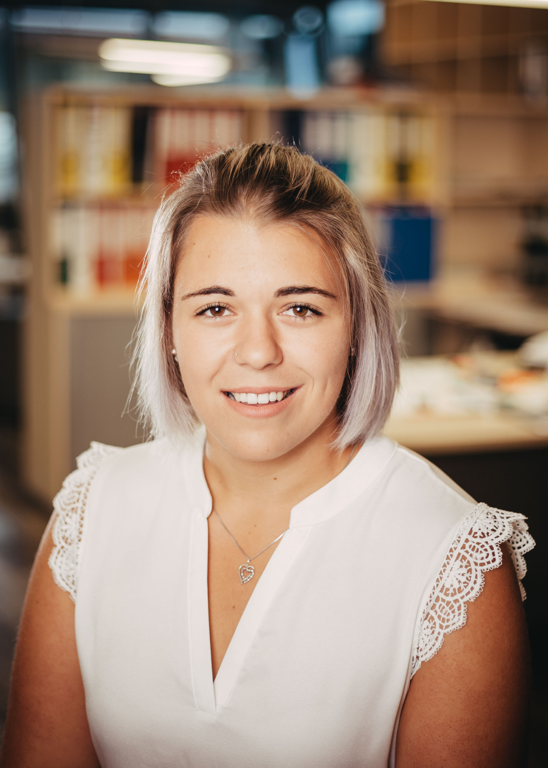 Porträt einer lächelnden Frau mit kurzem hellem Haar und weißer Bluse. Sie sitzt in einem modernen Büro, im Hintergrund sind Regale und ein unscharfer Arbeitsbereich zu sehen.
