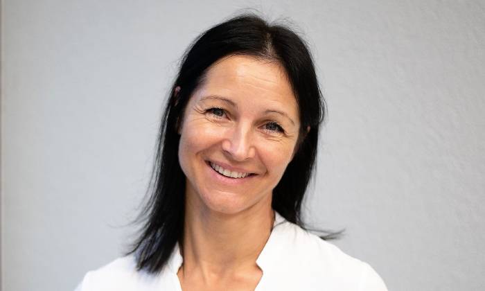 Das Bild zeigt eine Person mit schwarzem mittellangem Haar, wobei das Gesicht verpixelt ist. Die Person trägt ein schlichtes weißes Oberteil und steht vor einem grauen Hintergrund.