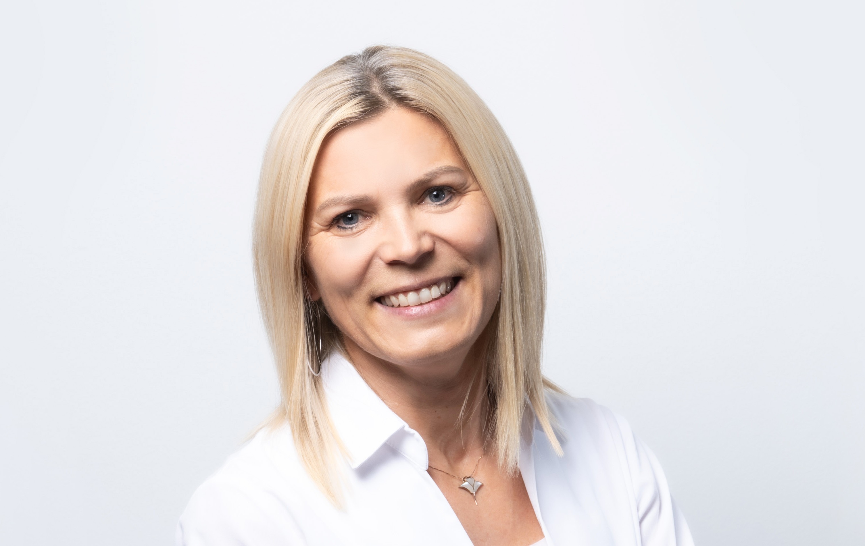 Freundliches Porträt einer blondhaarigen Frau in weißer Bluse vor hellem, neutralem Hintergrund. Sie lächelt direkt in die Kamera, das Bild wirkt professionell, sauber und natürlich.