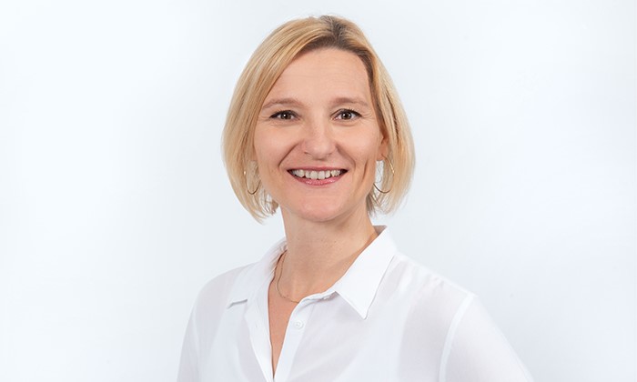 Eine Person trägt ein weißes Hemd und steht vor einem neutralen, hellen Hintergrund. Das Gesicht ist bewusst abgeblendet.